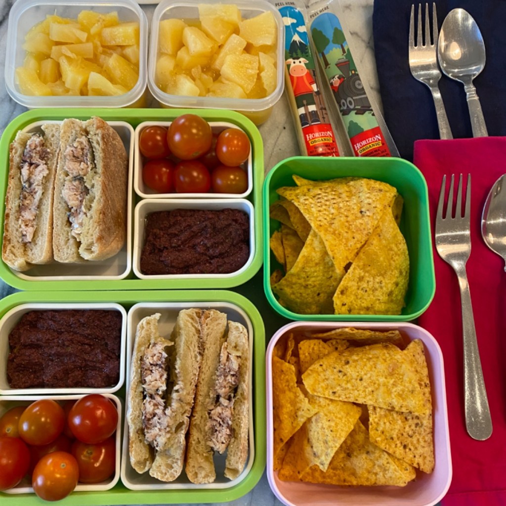 Homemade olive tapenade with cherry tomatoes and tortilla chips, along with sardines burgers, mozzarella cheese sticks, and pineapple chunks, in kids' lunch boxes