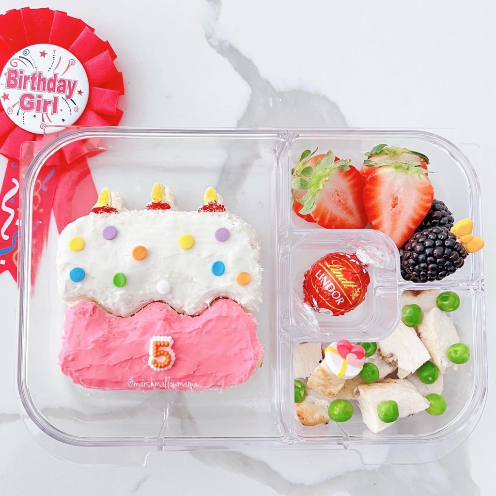 Birthday lunch packed in a kid's lunchbox, with cake-shaped pink sandwich, and other treats