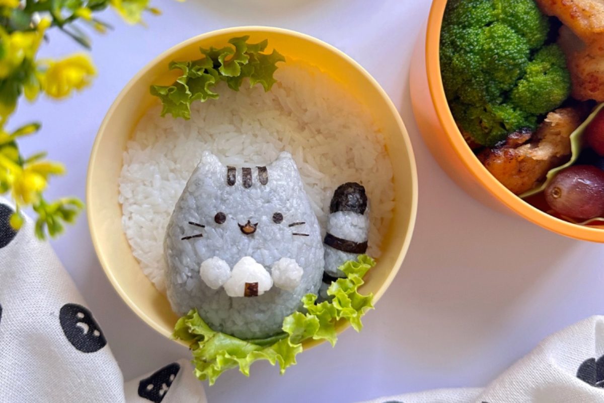 Cute cat-shaped onigiri rice ball in a colorful kids’ bento lunchbox with fresh vegetables and fruits.