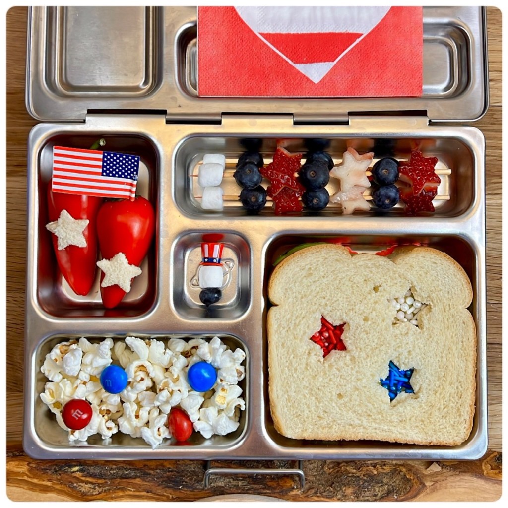 A festive Independence Day lunchbox featuring a sandwich with star-shaped cutouts, popcorn topped with red and blue M&Ms, and colorful skewers of strawberries, blueberries, and mini marshmallows. This red, white, and blue lunch idea is perfect for celebrating the Fourth of July with kids. Learn more and get inspired on Teuko.com.