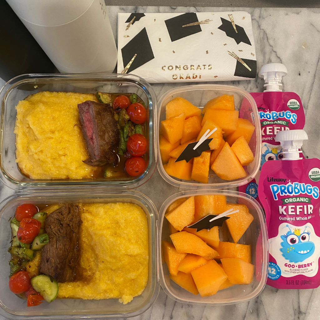 Meal prep lunchbox with Mediterranean-style beef steak, creamy polenta, zucchini and tomato Provençal, accompanied by kefir and diced melon for dessert, decorated with graduation hat food picks and napkins celebrating the end of the school year, packed for kids and teens school lunch by Teuko.
