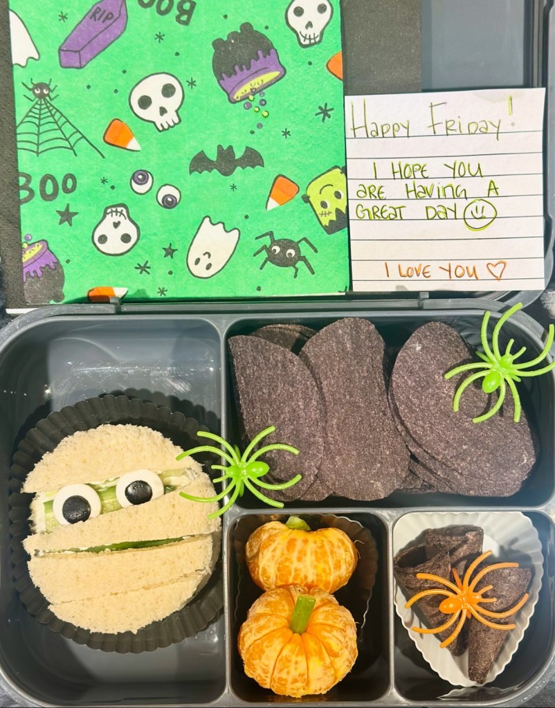 Handwritten note saying ‘Have a great day!’ inside a fun lunchbox with fruit and sandwiches.