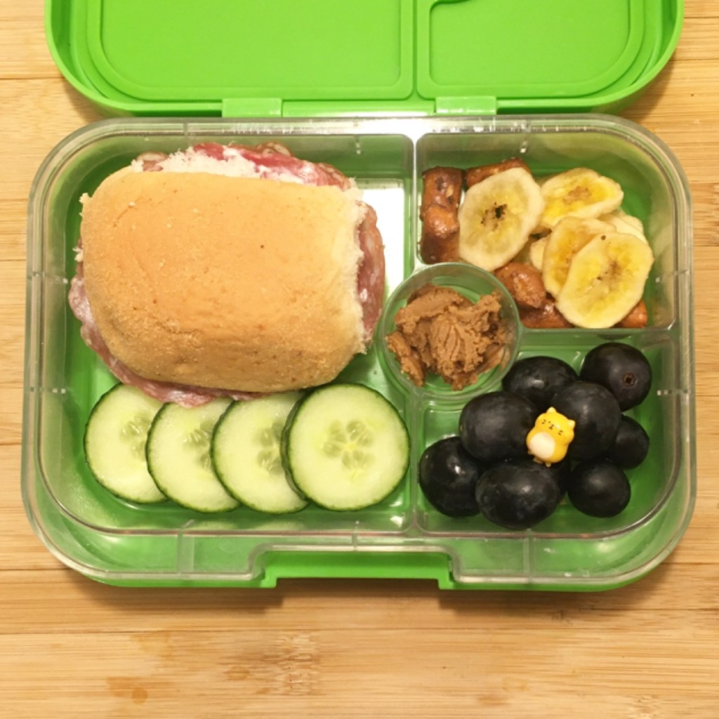 Banana chips with salami sandwich, cucumber slices, and blueberries, in a kids' lunchbox