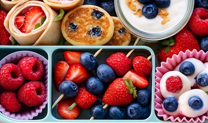 Overhead lunchbox with colorful berries, yogurt parfait, blueberry mini pancakes, and berry skewers in bright natural light.
