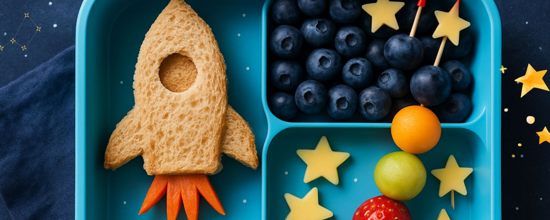 Space-themed lunchbox with a rocket-shaped sandwich, blueberries arranged like a galaxy, star-shaped cheese pieces, and colorful fruit skewers in a blue bento box on a starry background.