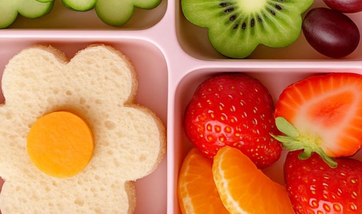 Spring-themed lunchbox with a flower-shaped sandwich, cucumber slices arranged as flowers, kiwi flower, strawberries, mandarin slices, and grapes in a pastel bento box.