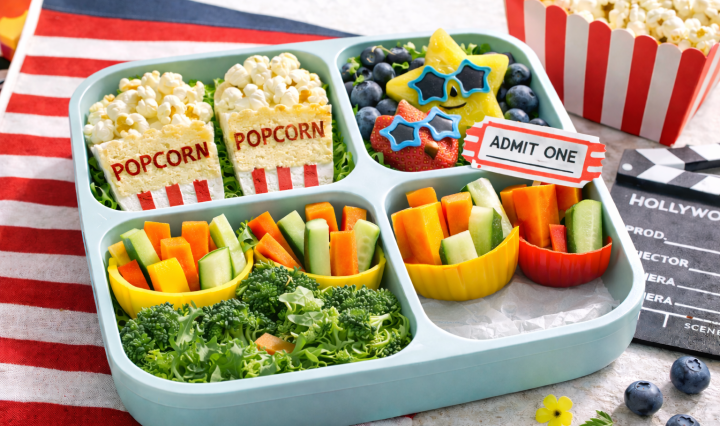 Movie-themed lunchbox with popcorn snacks, colorful finger foods, fruit art, and a lunchbox ticket note in bright natural light.