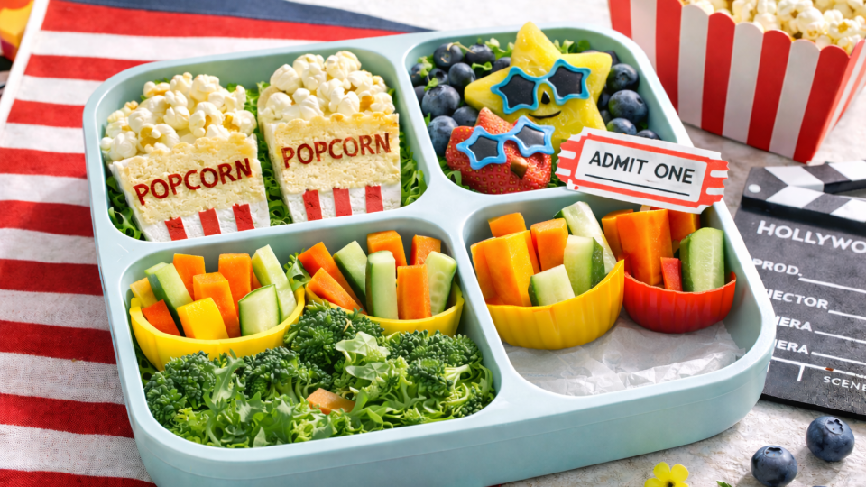 Movie-themed lunchbox with popcorn snacks, colorful finger foods, fruit art, and a lunchbox ticket note in bright natural light.
