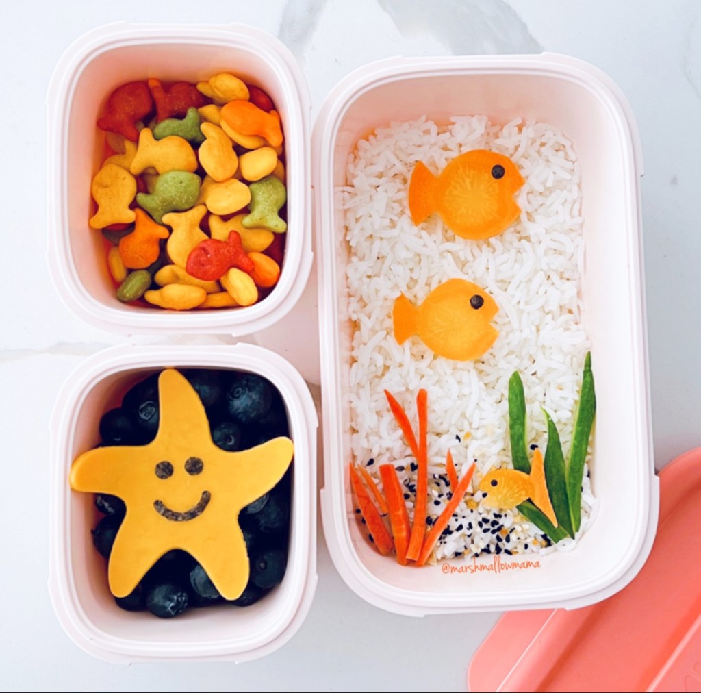 Ocean-themed lunchbox idea with colorful fish crackers, starfish slcied cheese, and fish-shaped carrot slices on top of white rice.