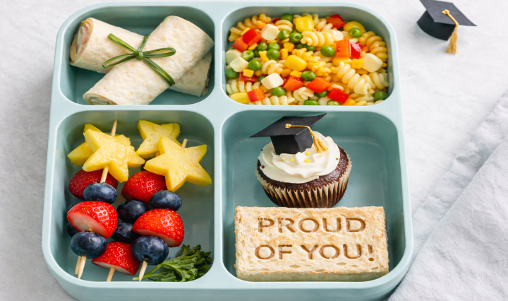 Graduation-themed bento lunchbox with tortilla diploma wraps, confetti pasta salad, star fruit skewers, and a cupcake topped with a graduation cap.
