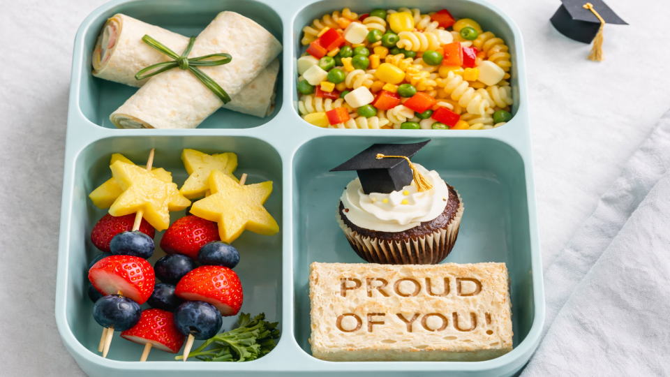 Graduation-themed bento lunchbox with tortilla diploma wraps, confetti pasta salad, star fruit skewers, and a cupcake topped with a graduation cap.