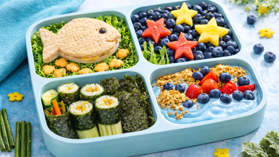 Ocean-themed lunchbox with fish-shaped sandwich, blueberry ocean, seaweed snacks, and star-shaped fruit in bright natural light.