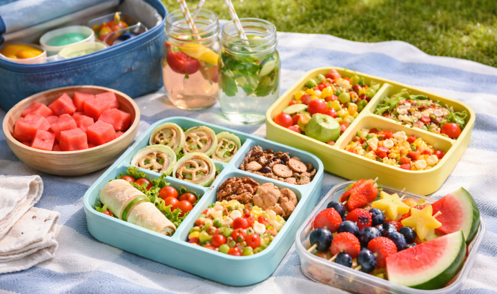 Picnic blanket with colorful lunchboxes, fruit skewers, wraps, pasta salad jars, and watermelon slices on a sunny day.