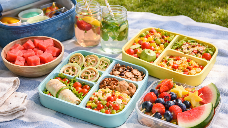 Picnic blanket with colorful lunchboxes, fruit skewers, wraps, pasta salad jars, and watermelon slices on a sunny day.